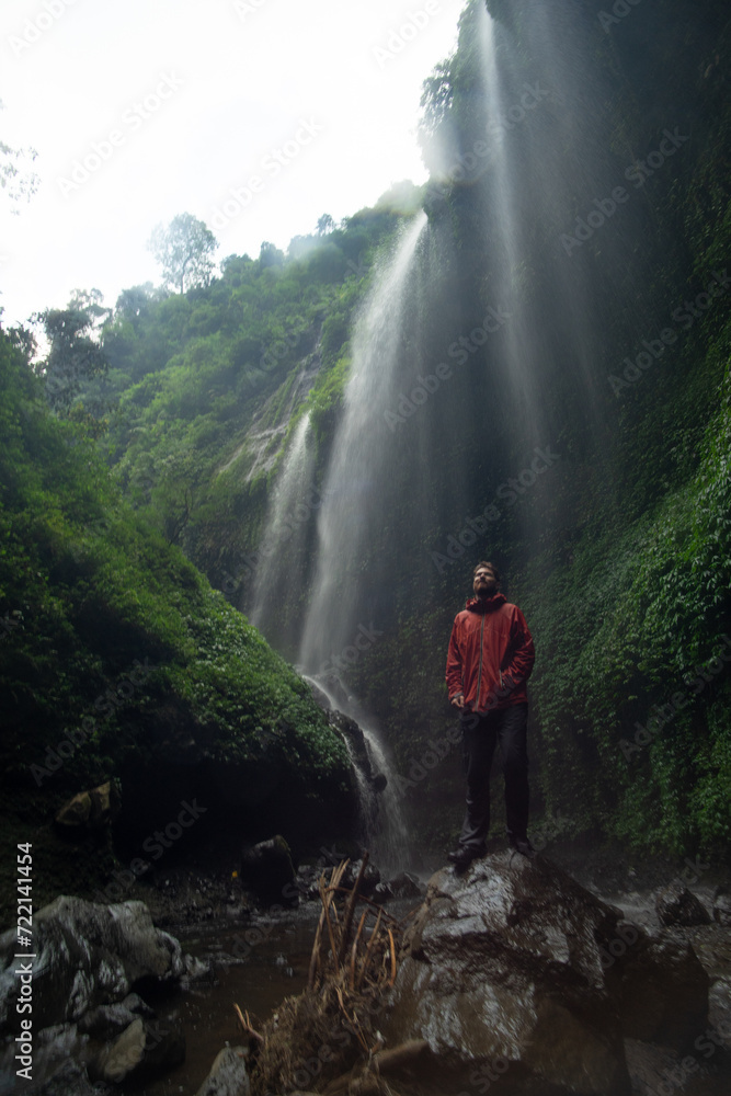 Person standing in front of a majestic waterfall surrounded by lush greenery Person standing in front of a majestic waterfall surrounded by lush greenery