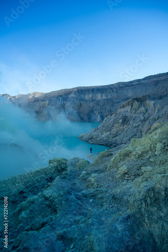 A lone figure stands near the misty waters of a volcanic crater lake surrounded by rugged terrain.