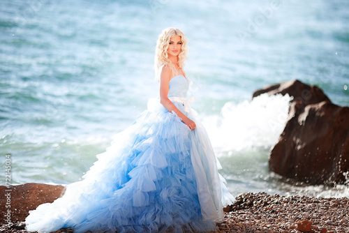 Beautiful young girl in a long blue lush dress is standing on the beach