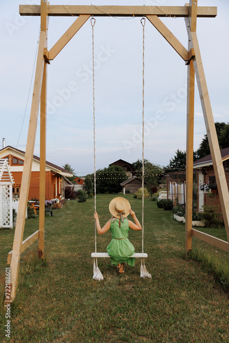 A Caucasian blonde woman in a flowing green dress enjoys a peaceful moment on a white swing in a lush garden. The concept of recreation, leisure and rural life outside the city.