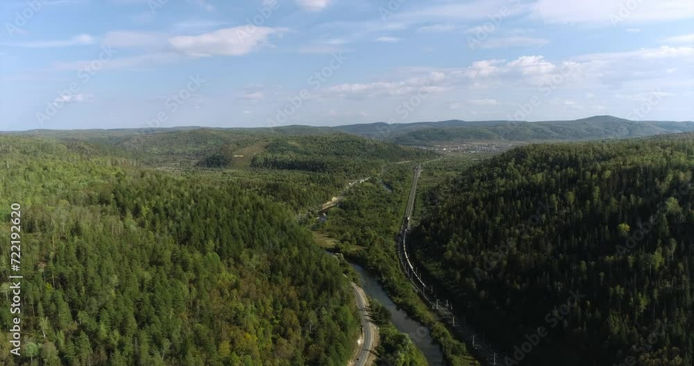 Freight long train carries with oil tank and petrol carriages an electric locomotive by Trans Siberian railways under the rock and near mountain river. Aerial drone wide view at summer sunny day Freight long train carries with oil tank and petrol carriages an electric locomotive by Trans Siberian railways under the rock and near mountain river. Aerial drone wide view at summer sunny day