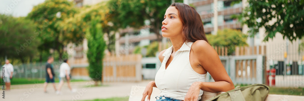 Lovely tanned woman with long brown hair wearing white top sitting relaxed on park bench looking away, Panorama. Life style concept Lovely tanned woman with long brown hair wearing white top sitting relaxed on park bench looking away, Panorama. Life style concept
