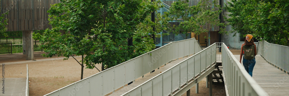 Fototapeta premium Back view, Cute tanned woman with long brown hair in white top and yellow bandana with backpack on her shoulders walks along the bridge, Panorama