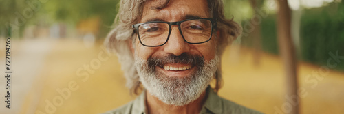 Clouse-up, friendly middle-aged man with gray hair and beard wearing casual clothes looks at the camera, Panorama. Mature gentleman in eyeglasses smiles at the camera