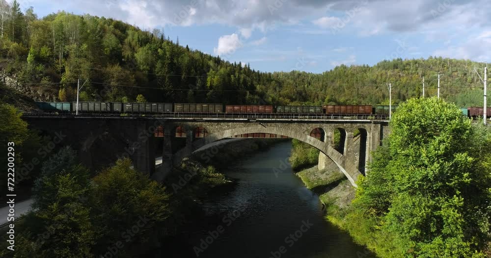 Freight long train carries with coal carriages on stone bridge by dangerous part of Trans Siberian railways near river in forest mountains. Aerial drone wide view at summer sunny sunset Freight long train carries with coal carriages on stone bridge by dangerous part of Trans Siberian railways near river in forest mountains. Aerial drone wide view at summer sunny sunset