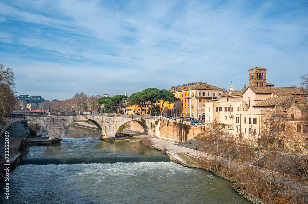Sunny winter day in Rome, featuring the small Tiber Island and Ponte Cestio. The scene is adorned with a beautiful bridge, colorful buildings along the waterfront, historic Italian cityscape