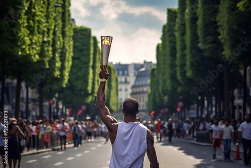 A male athlete in a white t-shirt holds a golden torch with a burning flame of the Sports Games in his hand raised above his head on the street of Paris on a sunny day.