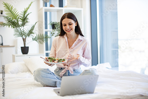 Front view of smiling woman having video chat with friends on laptop while eating healthy salad. Charming brunette resting on bed and enjoying nutrition from fresh vegetables while staying at home.