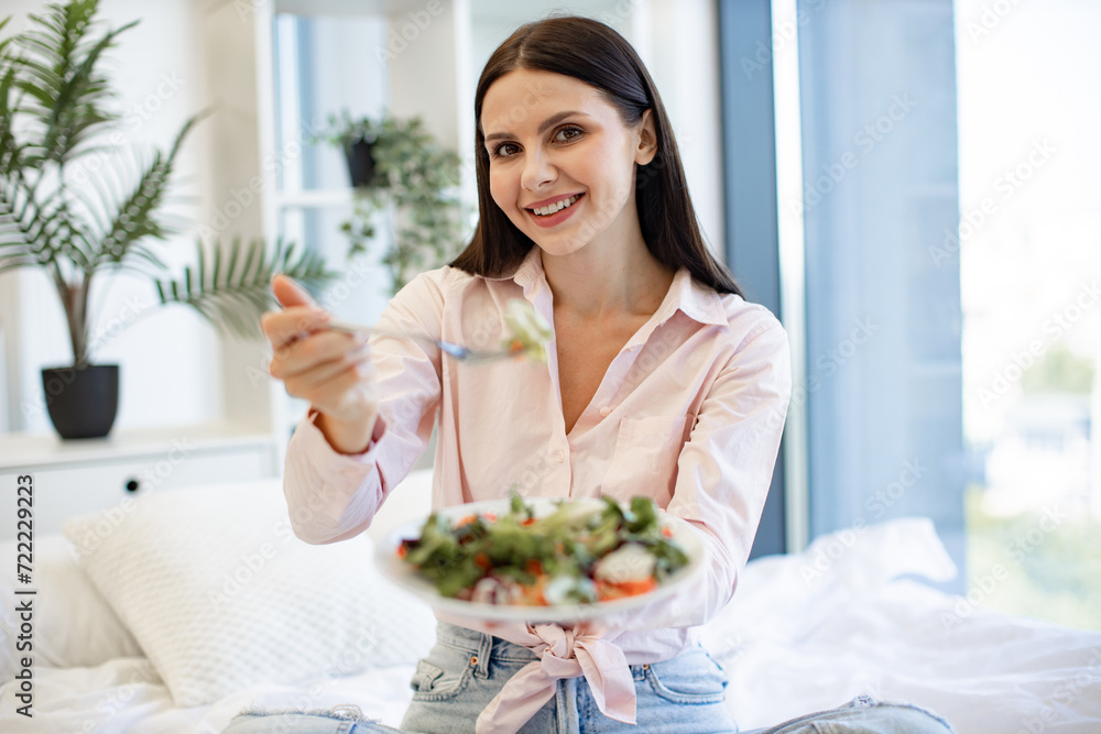 Portrait of brunette female enjoying breakfast from fresh vegetables on background of light modern cozy bedroom. Caucasian woman in casual wear sitting on comfy bed eating healthy salad.