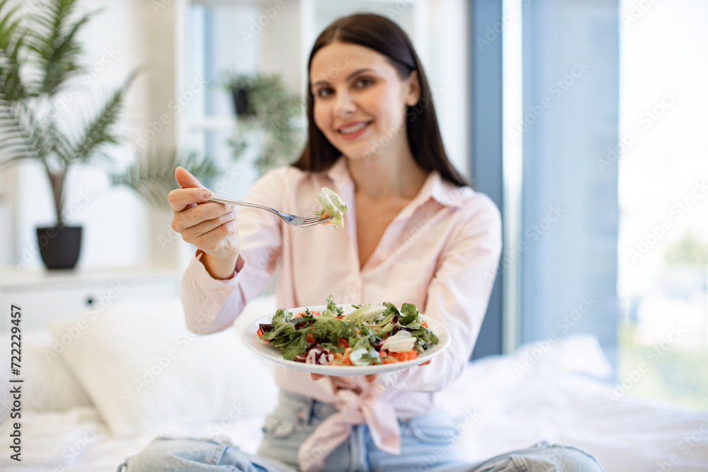 Focus on plate with salad and fork in hands of female enjoying breakfast in light modern cozy bedroom. Caucasian woman in casual wear sitting on comfy bed eating healthy salad.