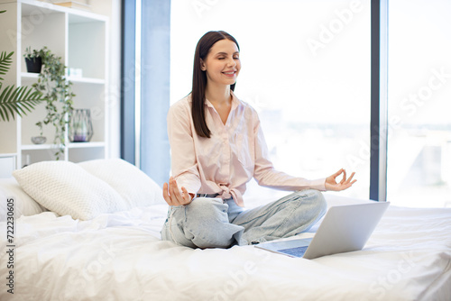 Caucasian woman enjoying practicing yoga during break with modern laptop at home. Peaceful lady sitting with closed eyes on bed on lotus position during online video class using modern laptop.