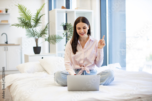 Portrait of young woman pointing finger up sign of having idea while working on modern laptop. Caucasian brunette in casual wear working remotely enjoying online chatting with colleagues from home .