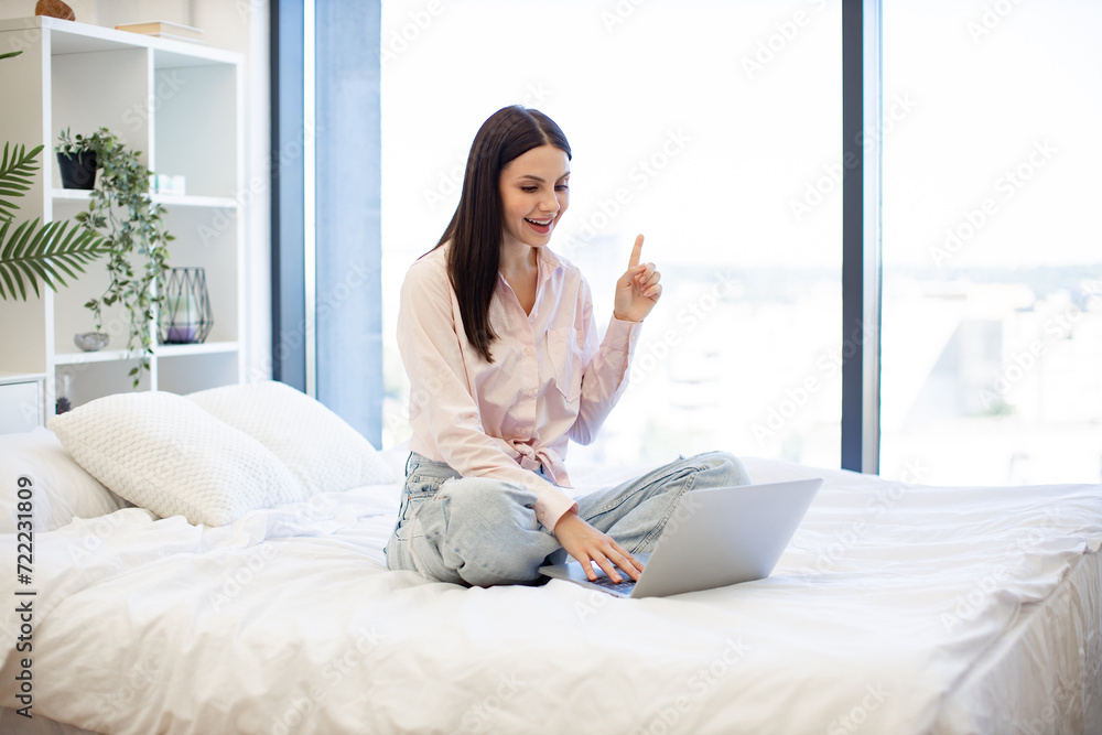 Portrait of young woman pointing finger up sign of having idea while working on modern laptop. Caucasian brunette in casual wear working remotely enjoying online chatting with colleagues from home . Portrait of young woman pointing finger up sign of having idea while working on modern laptop. Caucasian brunette in casual wear working remotely enjoying online chatting with colleagues from home .