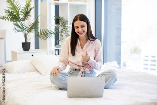 Front view of smiling woman having video chat with friends on laptop while sitting on comfortable bed showing thumb up. Charming female enjoying communication at distance using modern technologies.