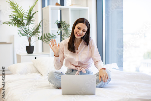 Happy brunette female waving hello while talking by video call using modern laptop, staying at home. Charming woman in casual attire enjoying online conversation with family while sitting on cozy bed.