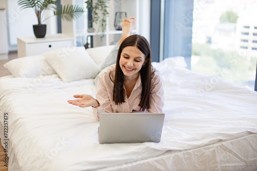 Charming woman in casual attire enjoying online conversation with friends while lying on cozy bed. Happy brunette female gesturing while talking by video call using modern laptop, staying at home.