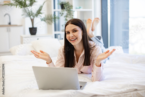 Front view of smiling woman having video chat with friends on laptop while lying on comfortable bed. Charming female enjoying communication at distance using modern technologies while staying home.