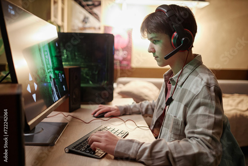 Side view portrait of teenage boy playing video games online on PC computer with wireless headset at night photo
