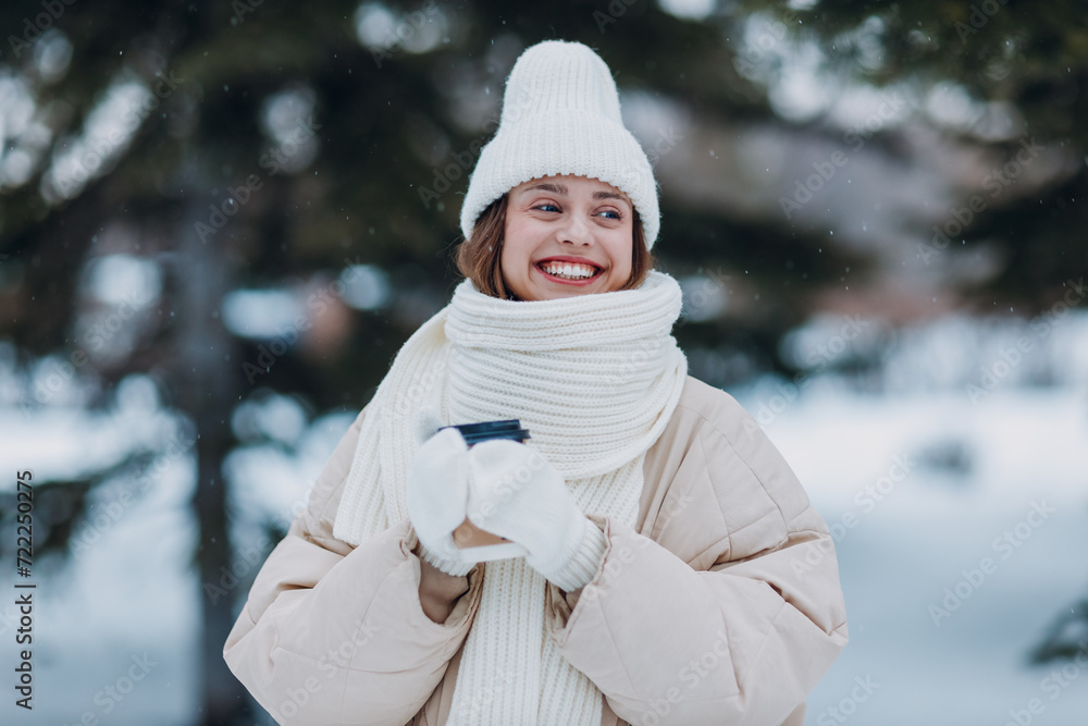 Obraz premium Young woman dressed coat scarf hat and mittens smiles and enjoys hot coffee at snowy winter park