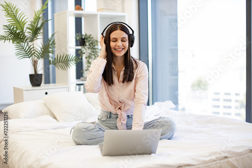 Creative young woman working remotely from home listening favorite music while typing on modern wireless laptop. Happy lady in headphones sitting on comfy bed and enjoying online chatting on weekend.