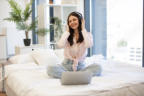 Smiling brunette female in wireless headphones enjoying favorite song, staying at home. Charming woman in casual attire dancing of favorite music during break of remote work while sitting on cozy bed.