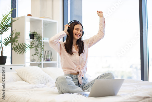 Beautiful young woman relaxing on cozy bed at home and using modern wireless headphones for listening positive songs. Caucasian brunette using modern devices enjoying favorite music on weekend.