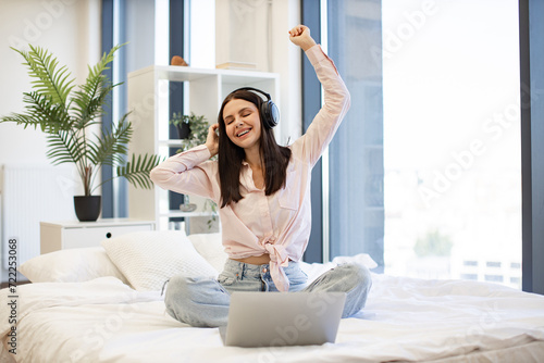 Caucasian brunette using modern devices enjoying favorite music on weekend. Beautiful young woman relaxing on cozy bed at home and using modern wireless headphones for listening positive songs.