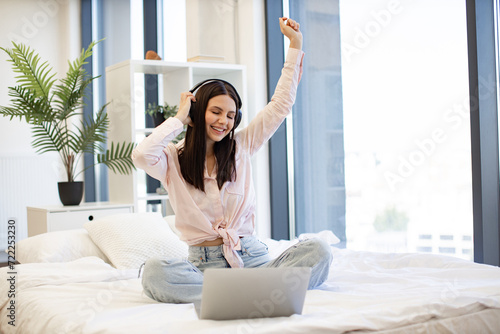 Caucasian brunette using modern devices enjoying favorite music on weekend. Beautiful young woman relaxing on cozy bed at home and using modern wireless headphones for listening positive songs.