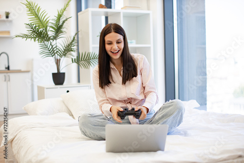 Happy young woman holding wireless joystick while playing video games on modern laptop. Happy lady sitting on comfy bed and enjoying favorite computer game on weekend.