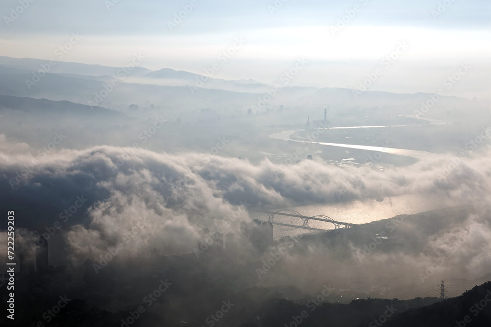 Naklejka premium Aerial panorama of Taipei City on a foggy morning with a bird's eye view of dense fog rolling over Guandu Plain, a bridge across Tamsui River & mountain silhouettes in the background