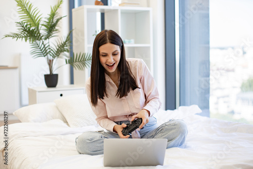 Beautiful young woman relaxing on cozy bed at home and using modern wireless joystick for playing games. Caucasian brunette using modern devices enjoying favorite video game on weekend.