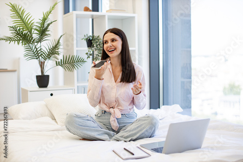 Charming woman in casual attire enjoying exchanging of audio messages while sitting on cozy bed. Positive caucasian female gesturing while recording conversation on modern smartphone, staying at home.