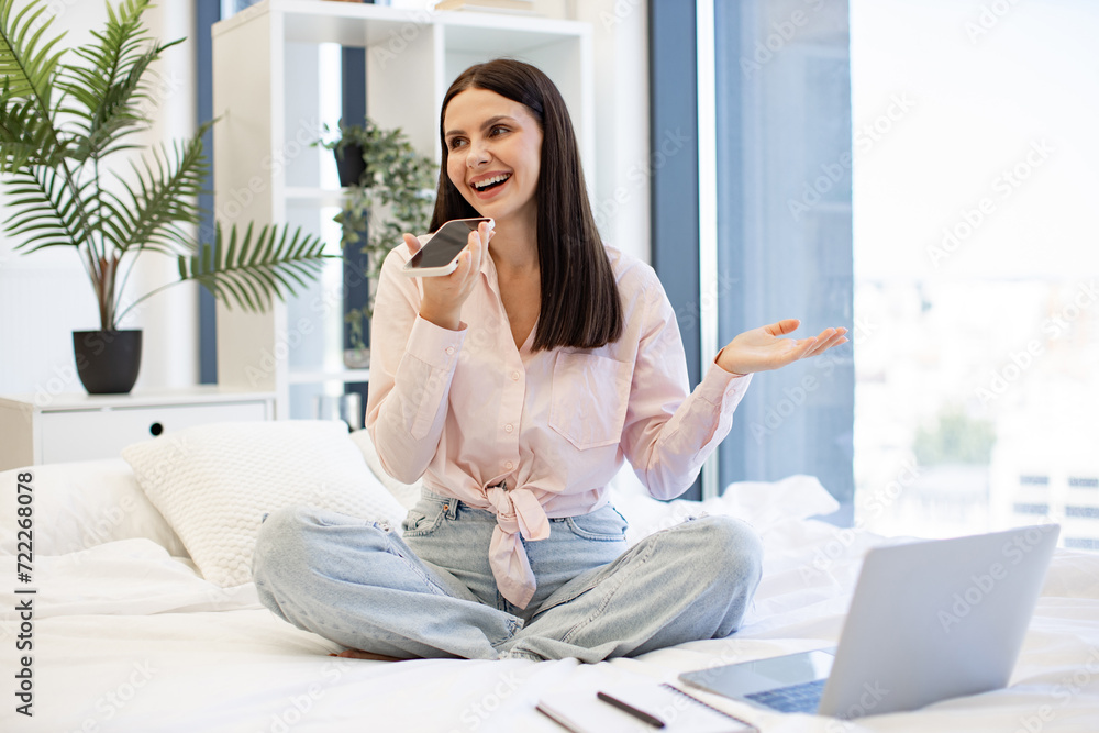 Charming woman in casual attire enjoying exchanging of audio messages while sitting on cozy bed. Positive caucasian female gesturing while recording conversation on modern smartphone, staying at home. Charming woman in casual attire enjoying exchanging of audio messages while sitting on cozy bed. Positive caucasian female gesturing while recording conversation on modern smartphone, staying at home.