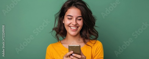 Young adult smiling happy pretty latin woman holding mobile phone looking at camera, doing ecommerce shopping on smartphone, using trendy dating apps on cellphone isolated on green background