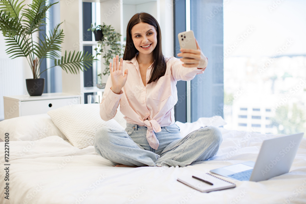 Beautiful lady sitting on comfy bed and enjoying online conversation with relatives or friends on weekend. Caucasian young woman holding wireless smartphone having a video call. Beautiful lady sitting on comfy bed and enjoying online conversation with relatives or friends on weekend. Caucasian young woman holding wireless smartphone having a video call.