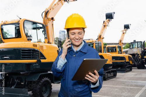 Engineer in a helmet with a digital tablet stands next to construction excavators