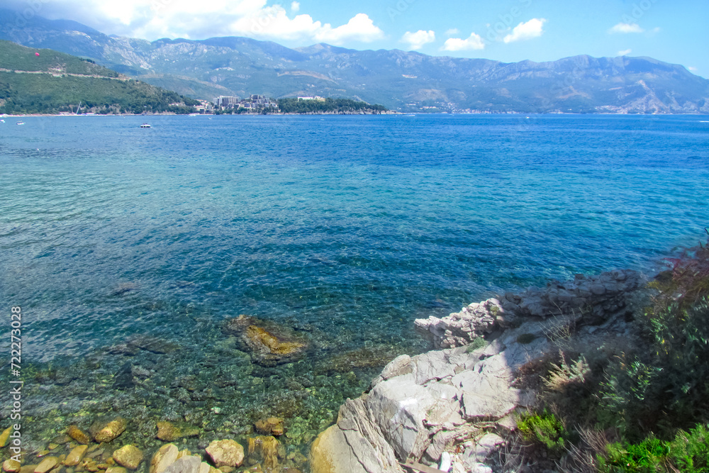 View of Adriatic Sea in sun's glare, shimmering with different shades from blue to turquoise and stones in clear water. A town in mountains in distance. Montenegro