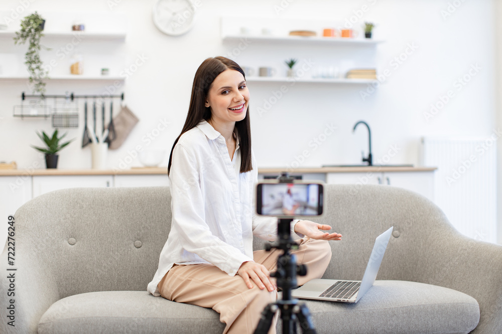 Front view of cheerful content maker in casual wear recording video to followers on modern smartphone. Young caucasian woman in with laptop sitting on comfortable sofa in kitchen.