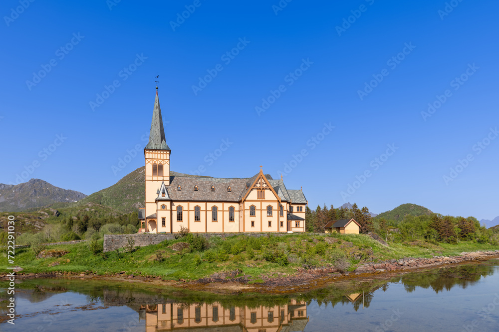 The Vagan Kirke in Lofoten, a stunning example of Gothic Revival architecture, reflects perfectly in the calm Norwegian waters under a vast blue sky