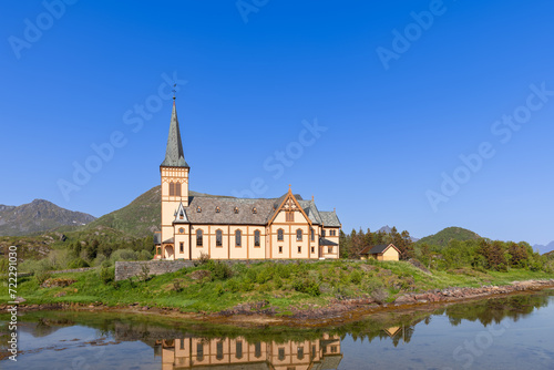 The Vagan Kirke in Lofoten, a stunning example of Gothic Revival architecture, reflects perfectly in the calm Norwegian waters under a vast blue sky © Artem