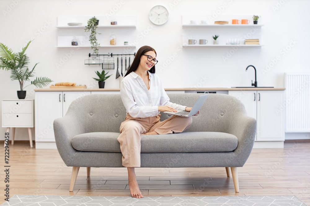 Obraz premium Concept of unification people and technology. Positive brunette young female in casual attire gesturing with hands while sitting on couch with modern laptop and looking at web camera.