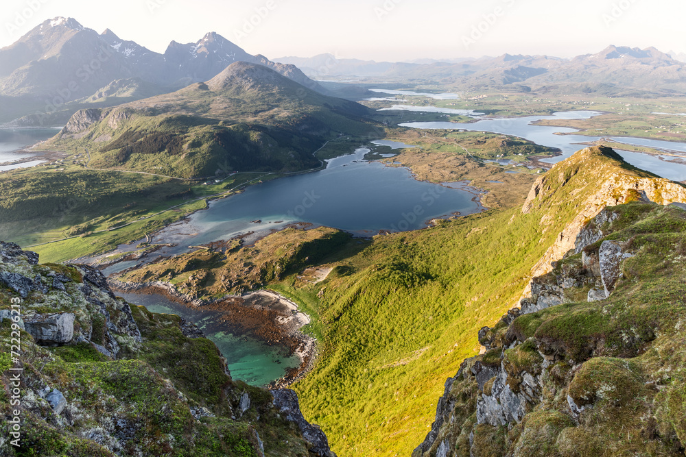 Golden sunlight bathes the verdant valleys and turquoise inlets of the Lofoten Islands, Norway, with a backdrop of majestic mountains, showcasing the raw beauty of the Arctic Circle