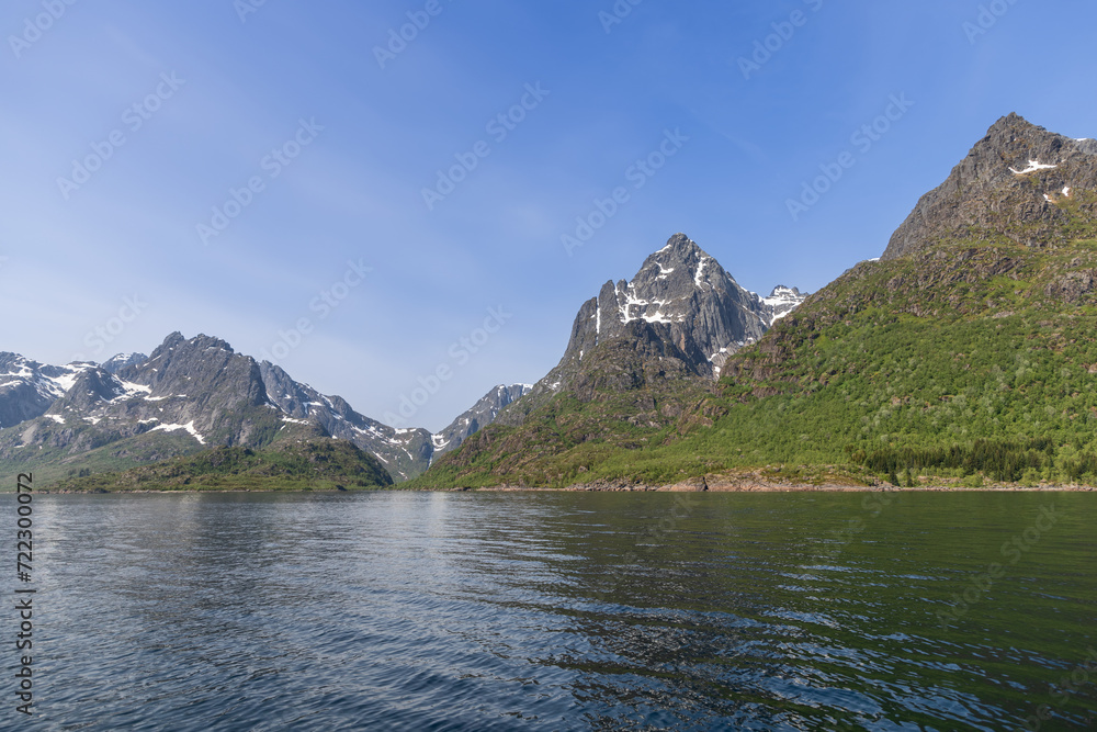 The stark Lofoten peaks, with lingering snow, stand tall above the glistening waters of a Norwegian fjord, embodying the vibrant essence of summer The stark Lofoten peaks, with lingering snow, stand tall above the glistening waters of a Norwegian fjord, embodying the vibrant essence of summer