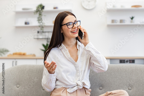 Positive caucasian female gesturing while having conversation on modern smartphone, staying at home. Charming woman in casual attire enjoying communication with colleagues while sitting on cozy sofa.