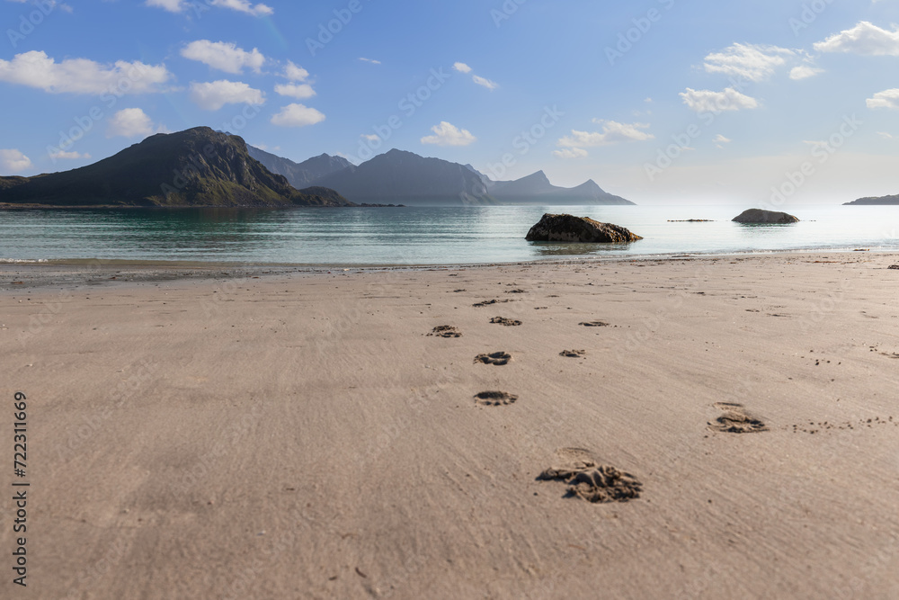 Footprints trail towards the sea on Haukland Beach, Lofoten, Norway, with rocky islets and misty mountains under a soft morning light (selective focus)