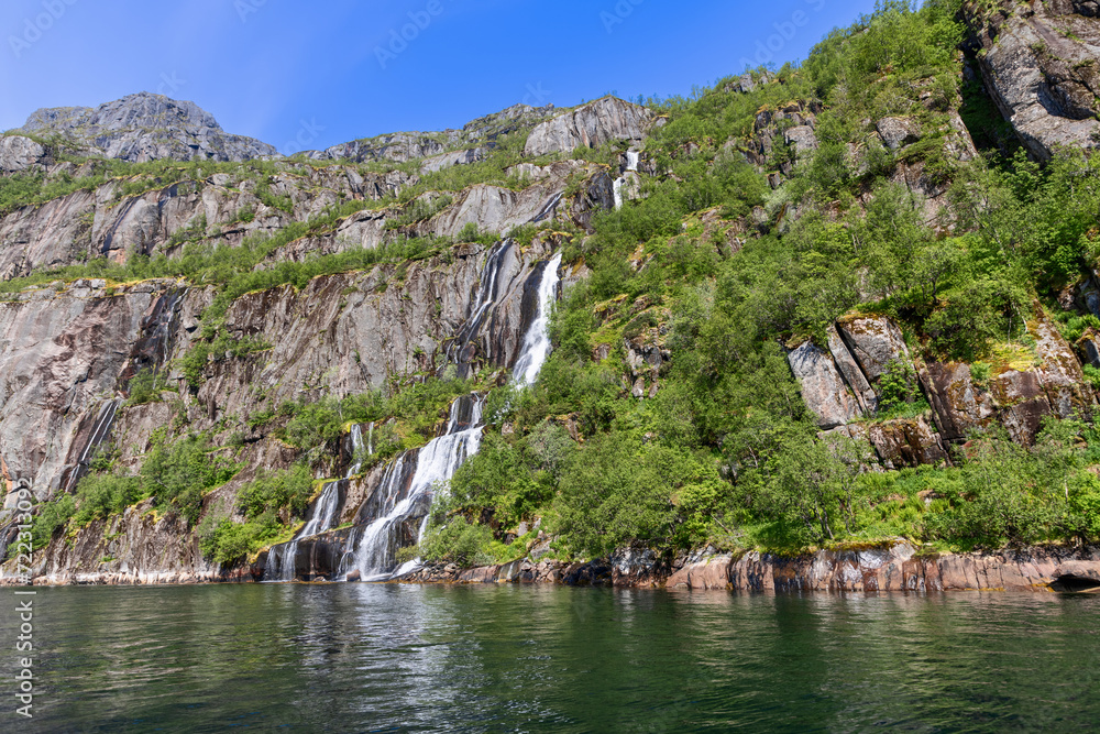 A bright summer day in Lofoten unveils a spectacular waterfall spilling over the cliffs into the clear waters of Trollfjorden, Norway. A bright summer day in Lofoten unveils a spectacular waterfall spilling over the cliffs into the clear waters of Trollfjorden, Norway.