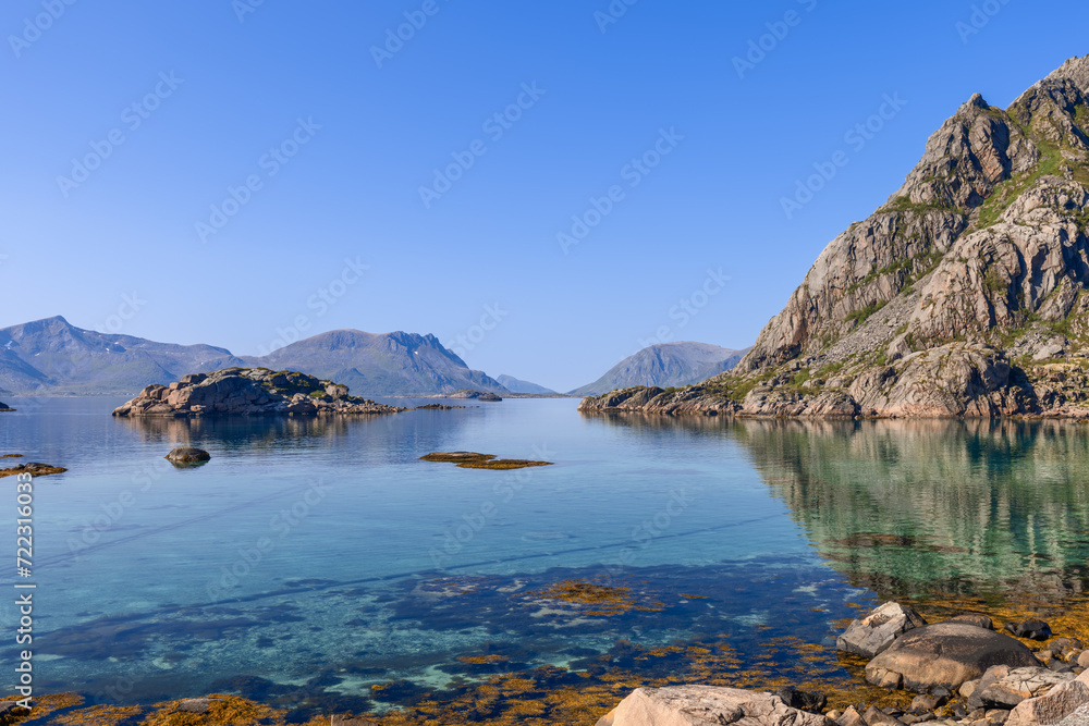 The calm waters of the Norwegian Sea gently lap against Lofoten's rocky coastline, with majestic mountains reflecting in the clear waters under a tranquil sky