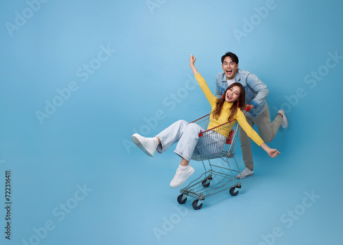 Happy fun Asian woman sitting inside of shopping trolley and man pushing shopping cart to get the latest offers promotion at the supermarket isolated on blue background.