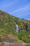 Vertical view of a cascading waterfall in Trollfjorden, Norway, cutting through lush greenery against a vibrant blue sky. Lofoten Islands, Norway