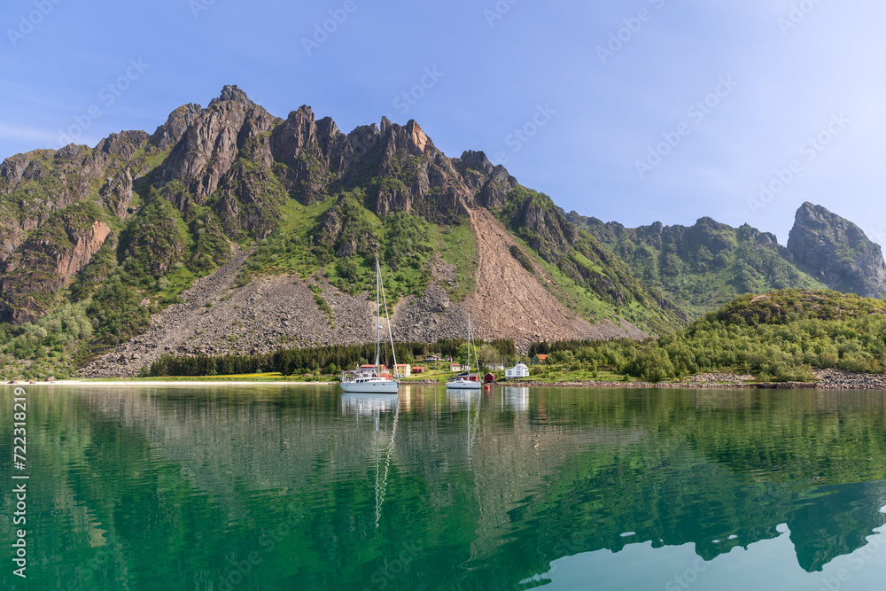 A tranquil summer scene in the Lofoten Islands, where sailboats and traditional Rorbu cabins are mirrored in the still waters under a rugged mountainous backdrop. Norway